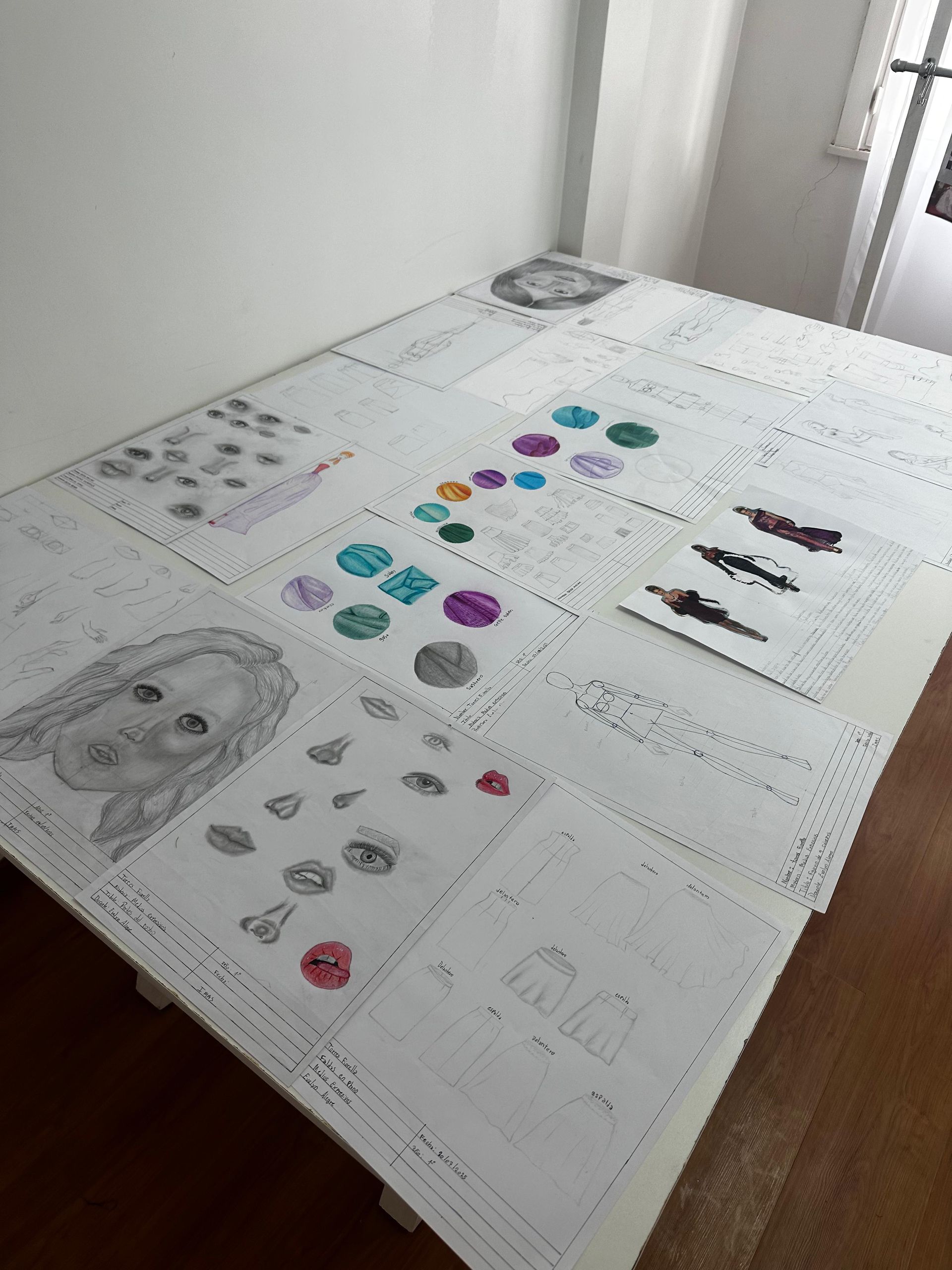 Sobre una mesa hay papeles de un proyecto artístico, incluyendo bocetos, muestras de color y un dibujo de un rostro.