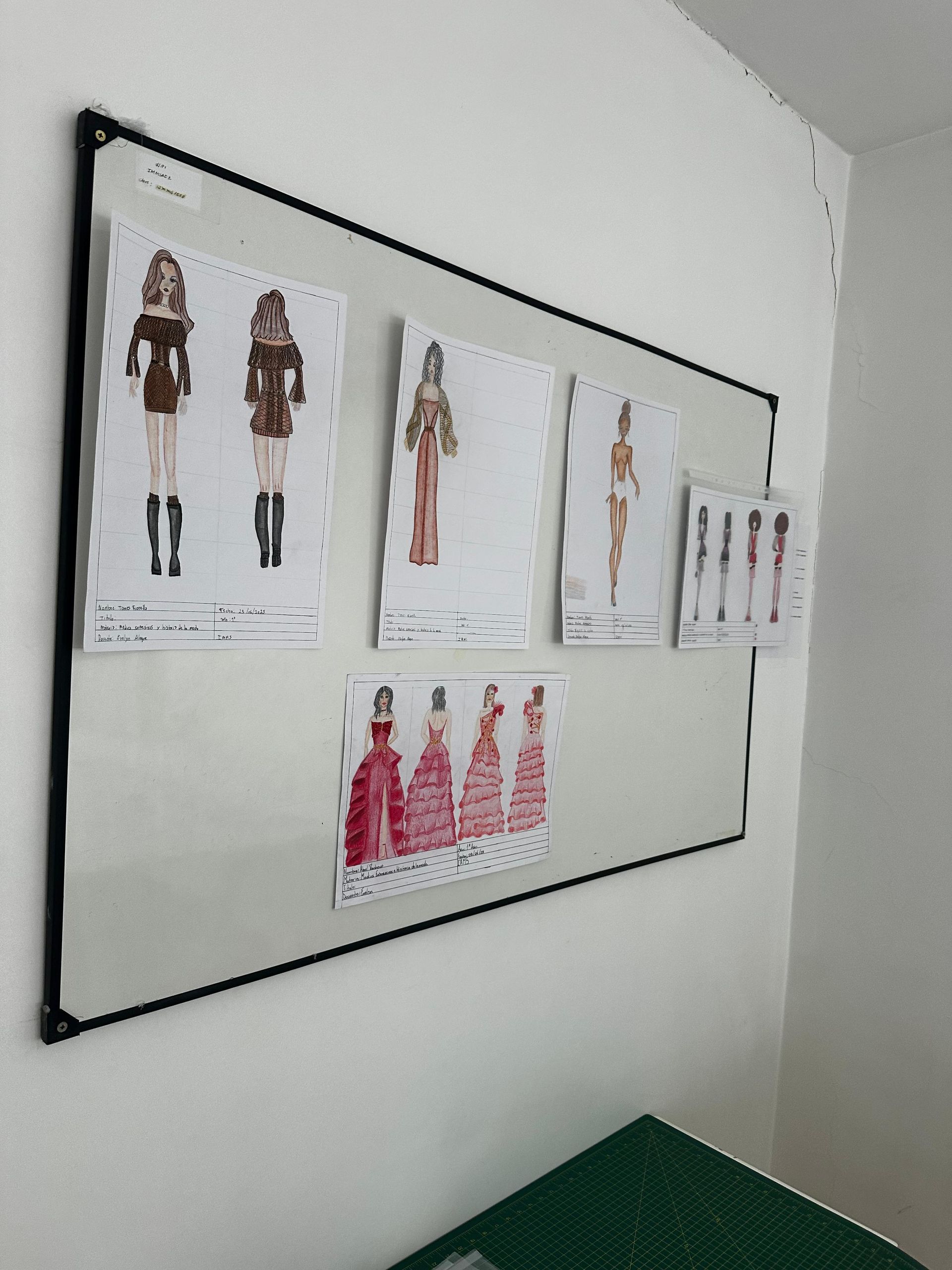 Bocetos de diseño de moda montados en la pared sobre una pizarra blanca encima de una mesa verde.