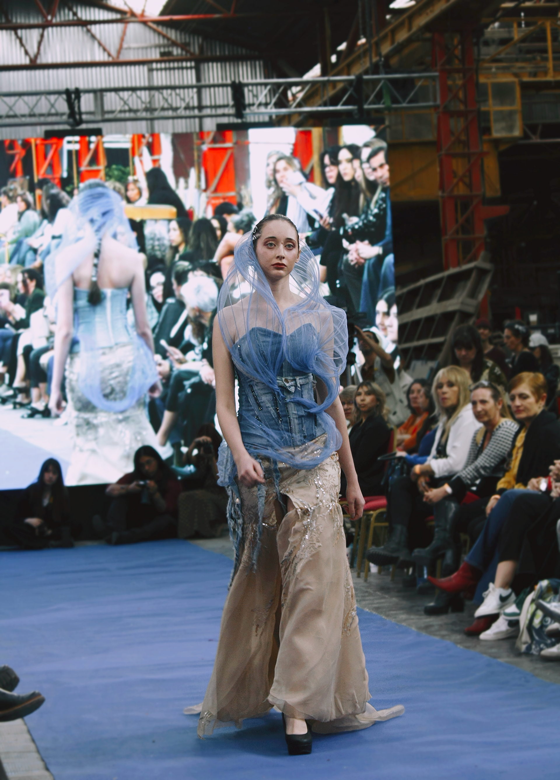 Una modelo con un conjunto azul y beige desfila por la pasarela ante un público sentado.