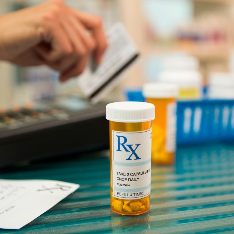 A pharmacy setting: prescription bottle on counter, hand holding a credit card over a register.