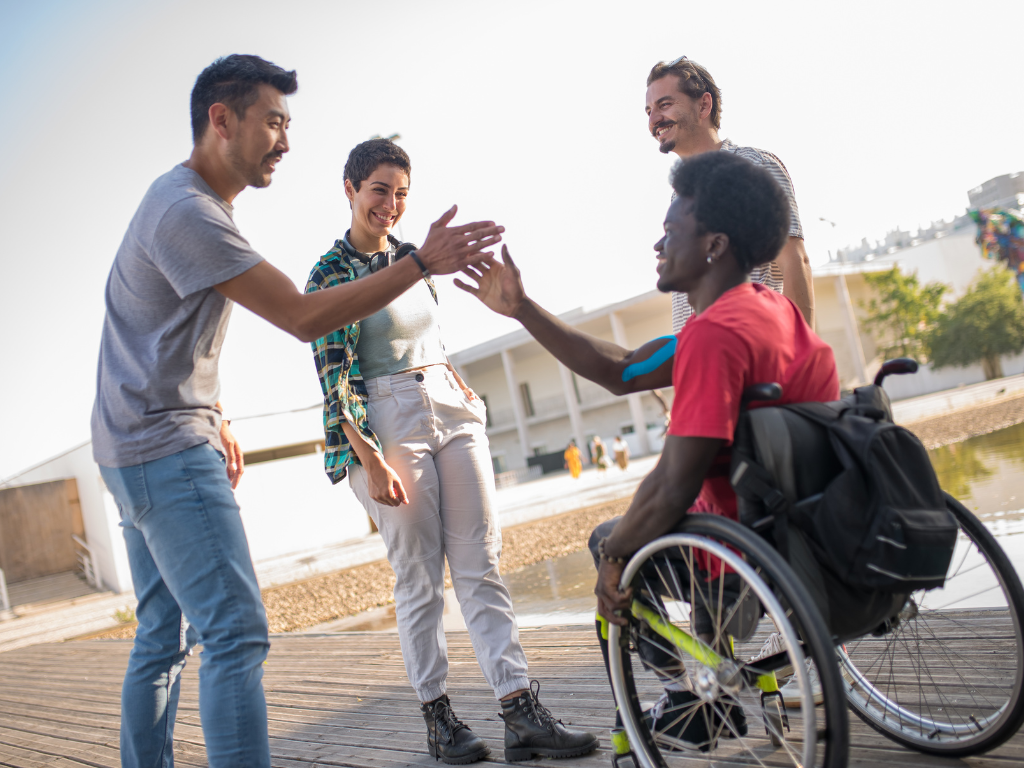Four diverse friends high-fiving outside near water; one in a wheelchair, smiles.