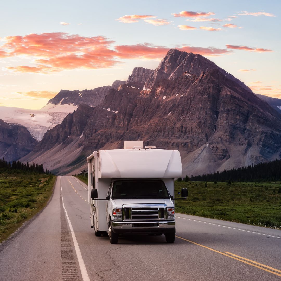 RV driving on an empty road toward mountains at sunset.