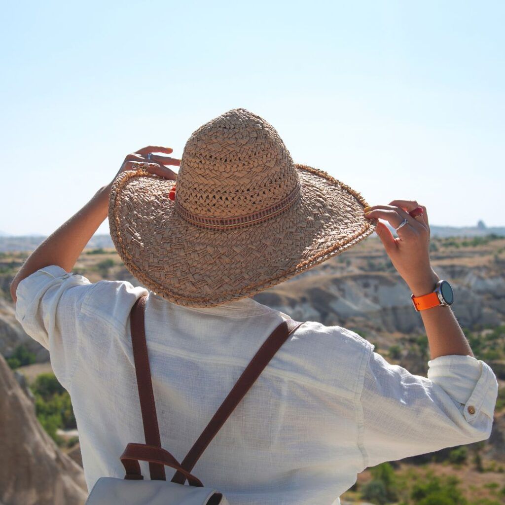 Person wearing a straw hat, looking at a scenic landscape. They have a backpack and watch on their arm.