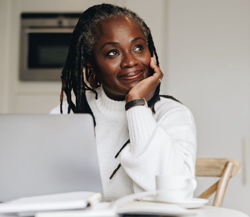 Person in a white sweater leaning on hand at a laptop, smiling thoughtfully at a table.