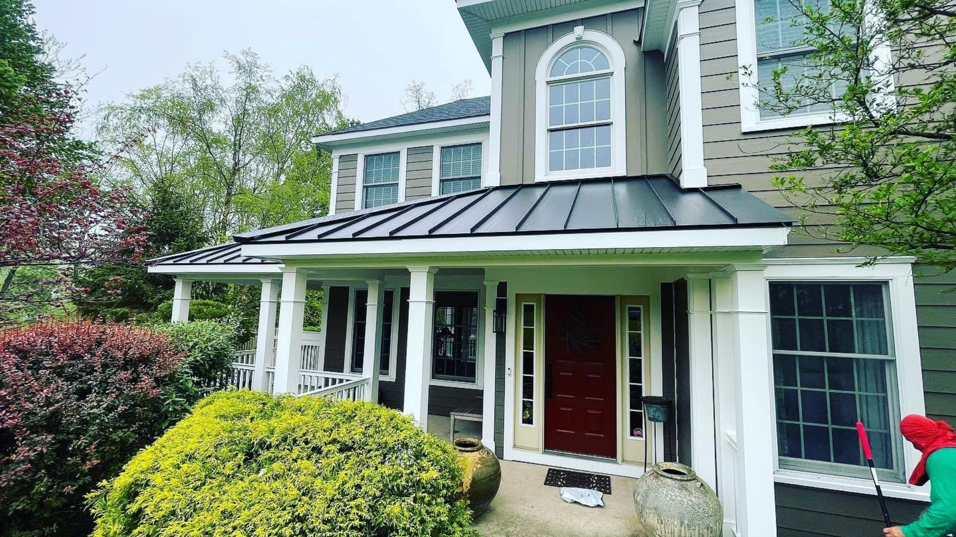 Two-story green house with white trim, red front door, and covered porch surrounded by lush landscaping (a project of Paese Construction LLC)
