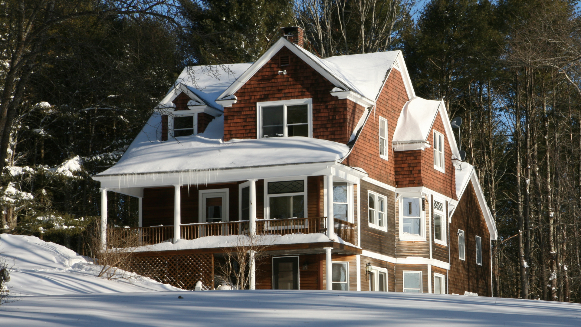 Snow-covered red brick house with white trim and wraparound porch in a winter forest