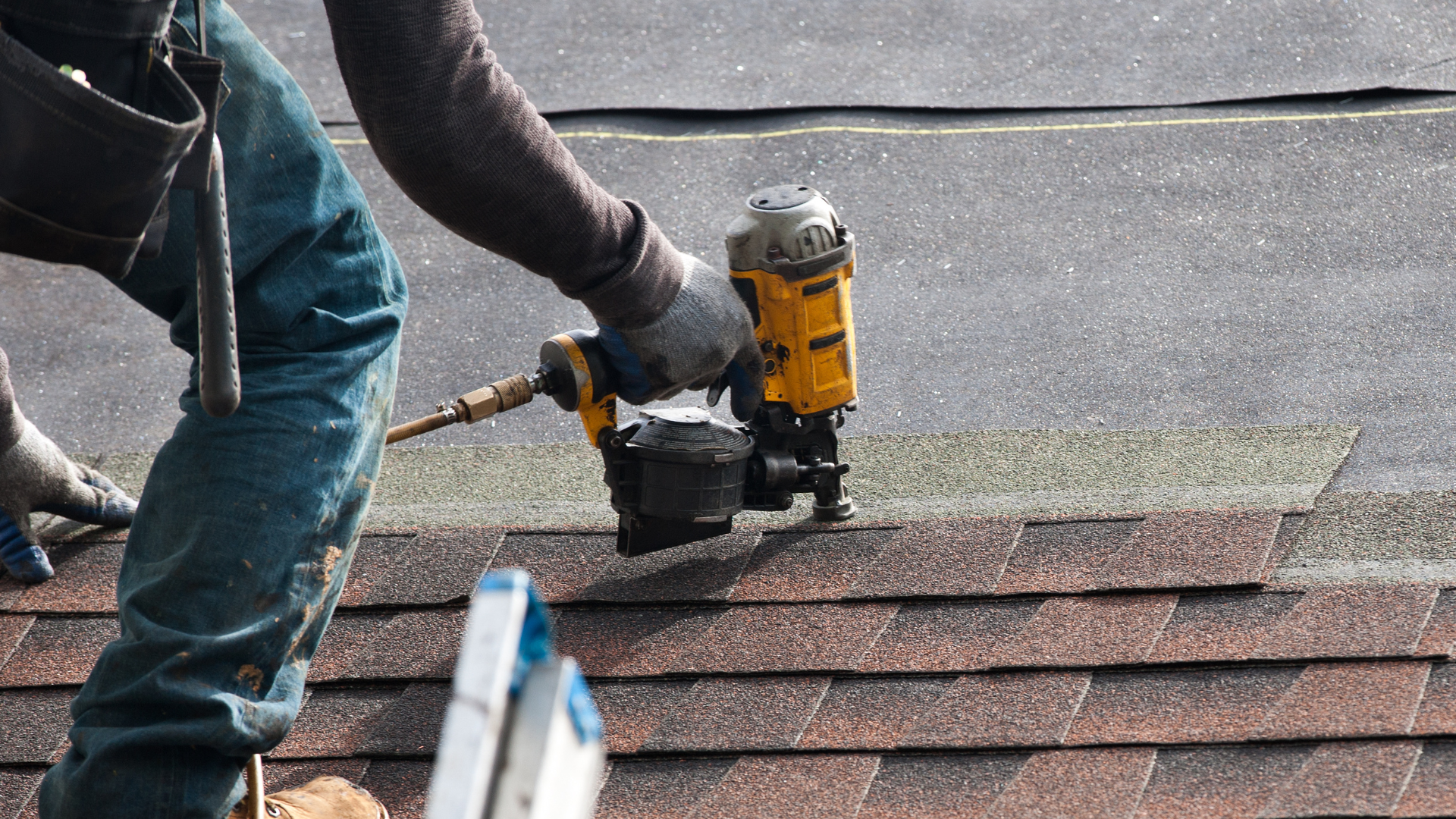 Worker using a yellow nail gun to install dark roof shingles on a sloped rooftop