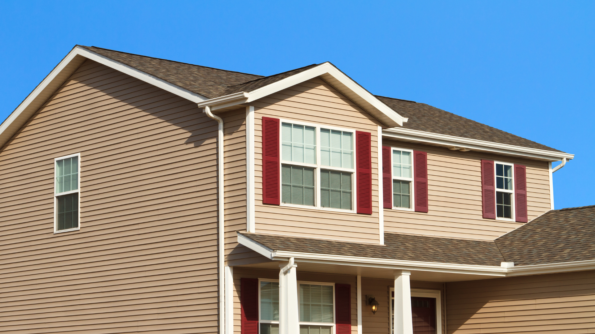 Suburban house exterior with beige siding, white trim, and red shutters under a blue sky