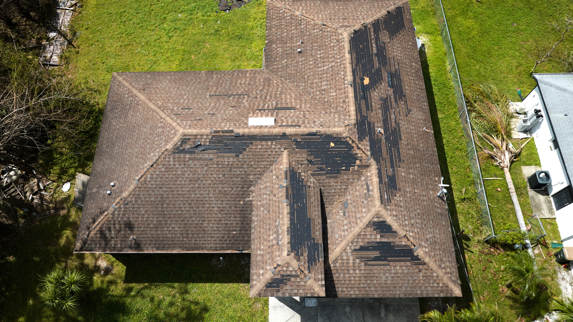 Aerial view of a brown-roofed house surrounded by green grass and a driveway with parked vehicles.