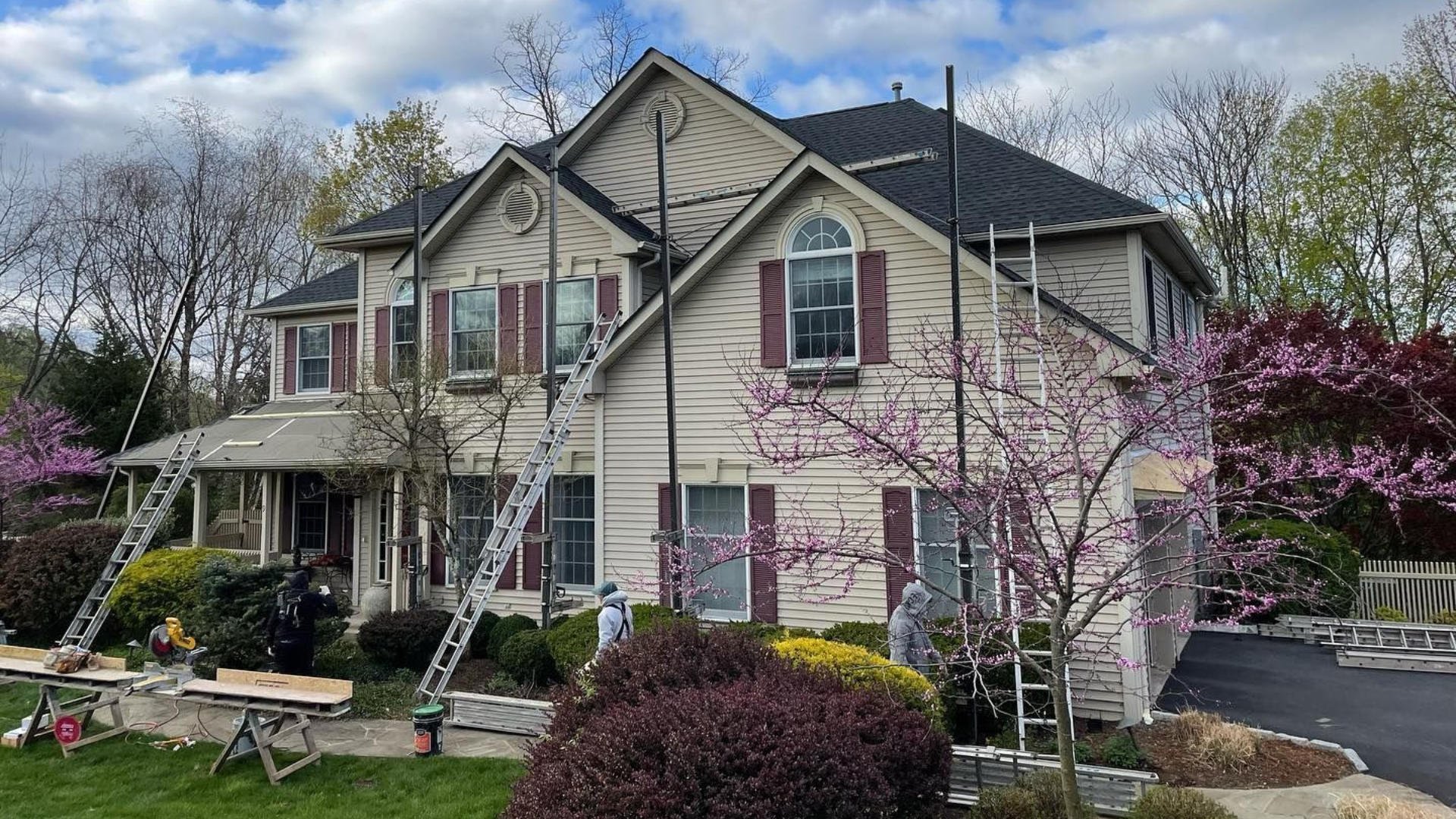 Two-story beige house with white trim, front porch, and blooming pink trees in spring