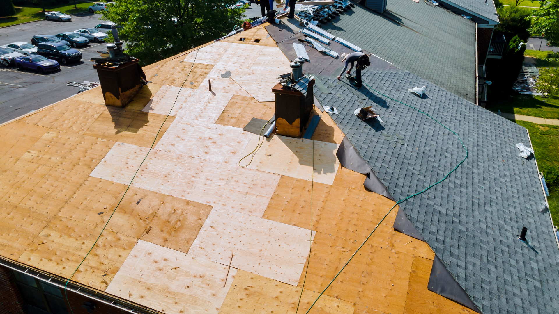 Roof under renovation with plywood sheathing and gray shingles, two workers on top near a residential street.