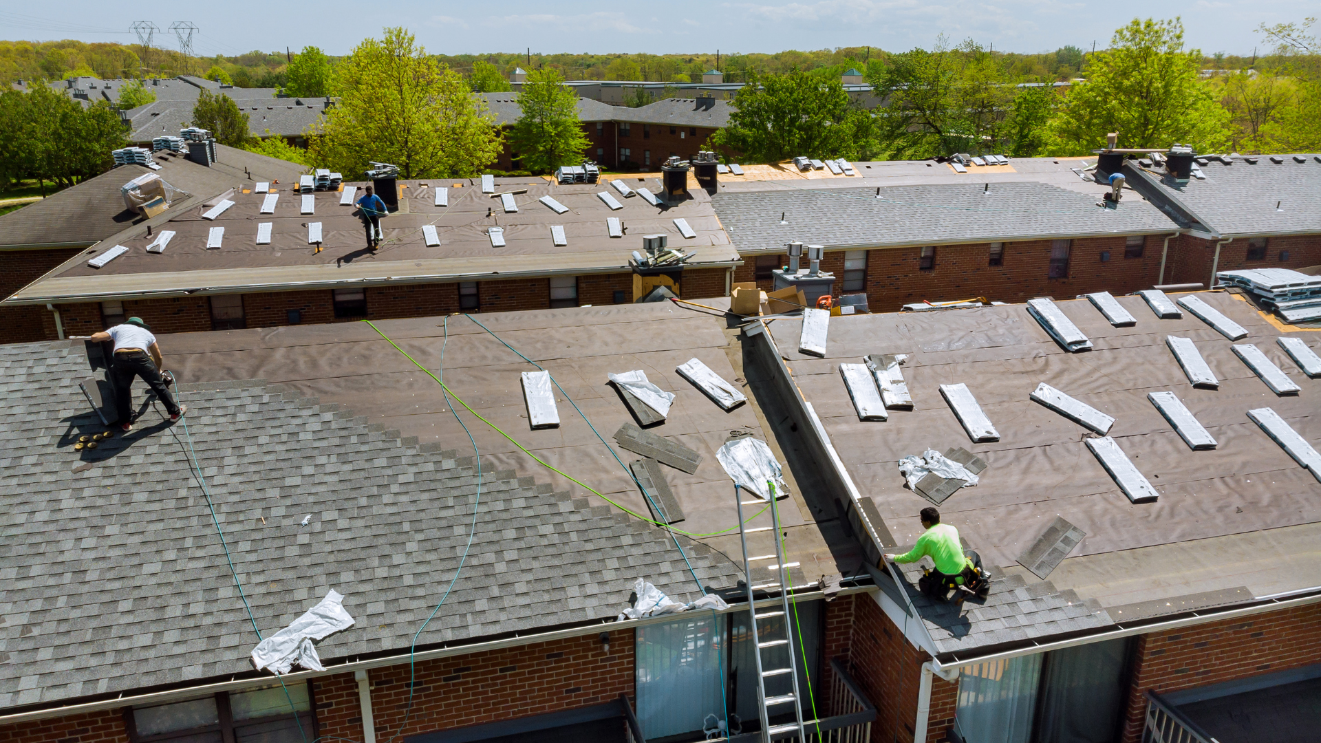 Roofing crew installing shingles on a large residential roof, with materials spread across multiple sections.