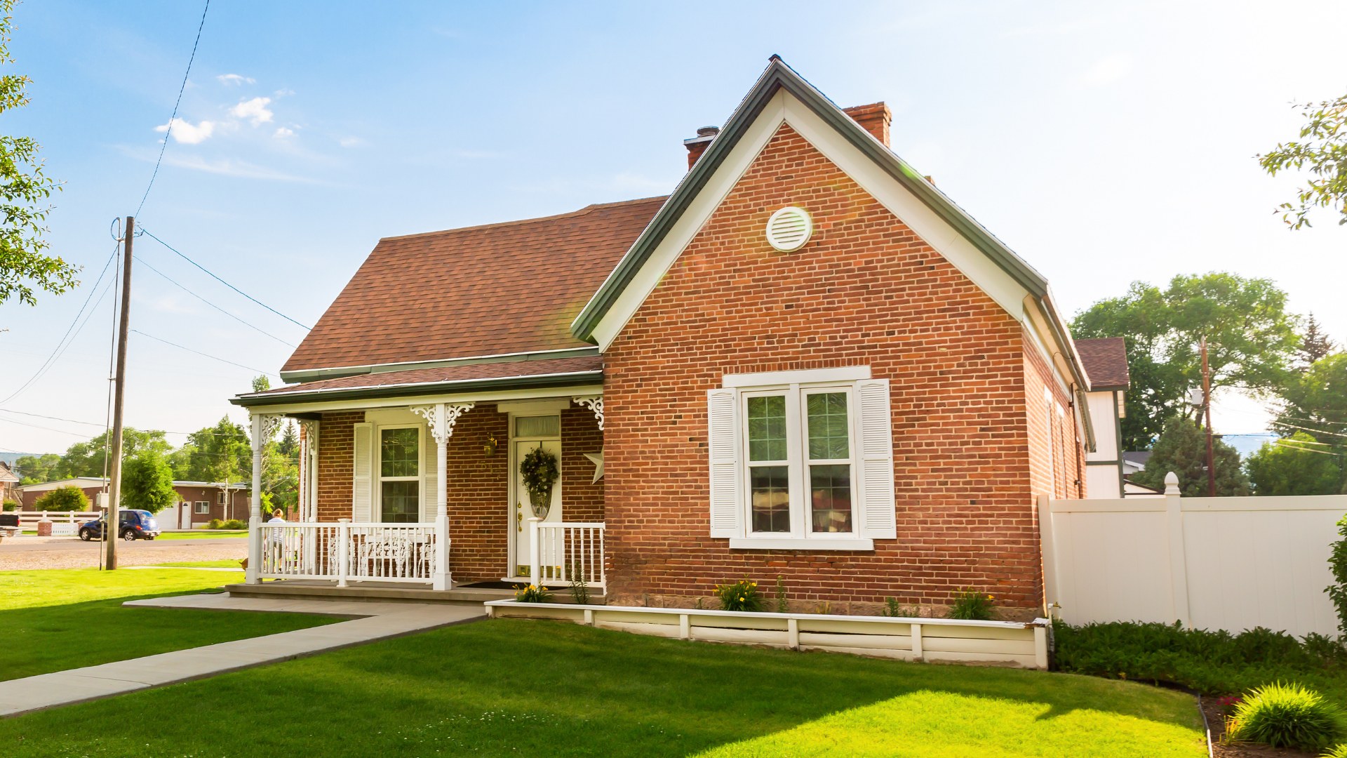 Brick house with white porch and shutters, red roof, and green lawn on a sunny day