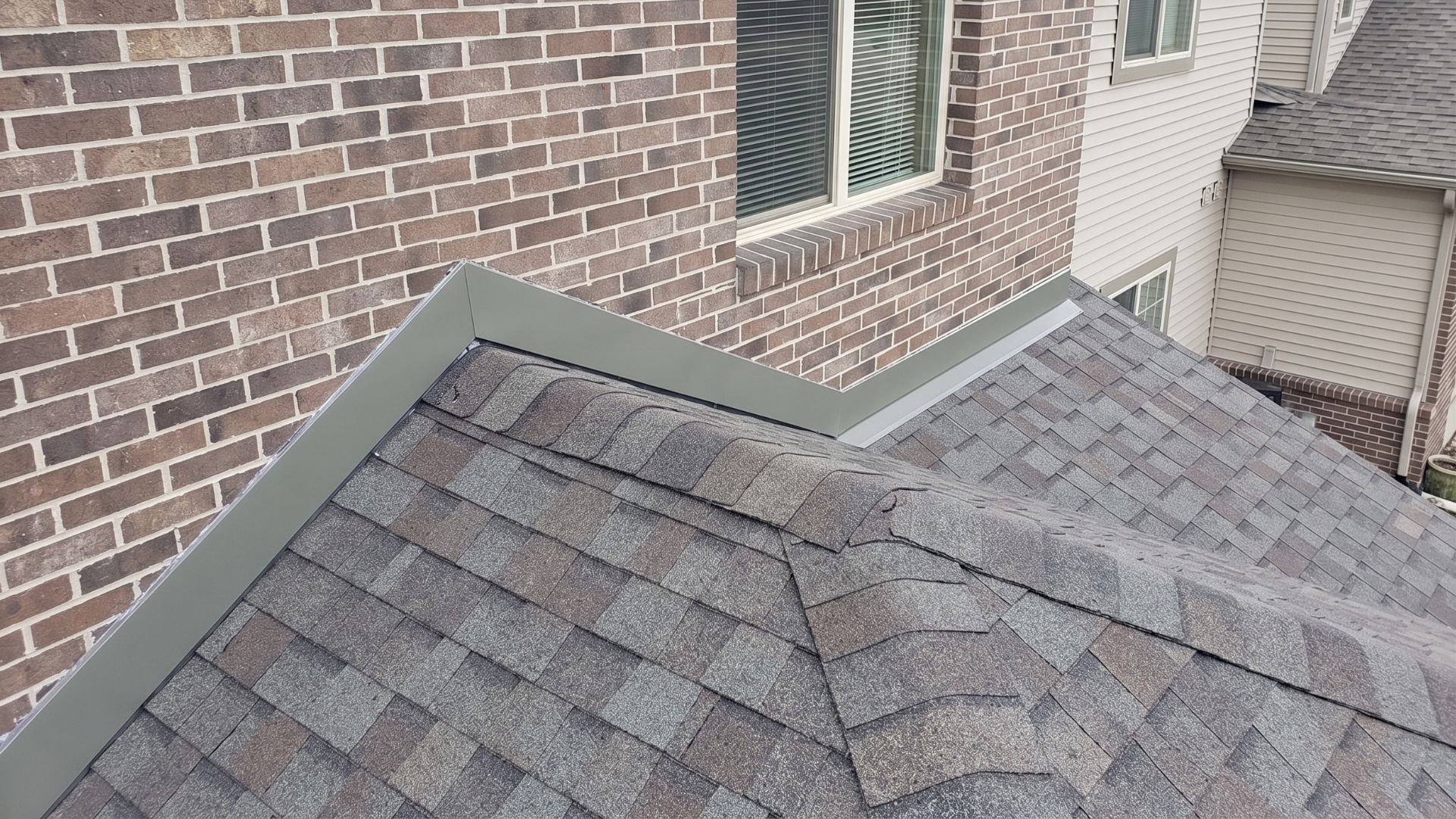 Corner of a shingled roof beside a brick house and neighboring siding with flashing in between