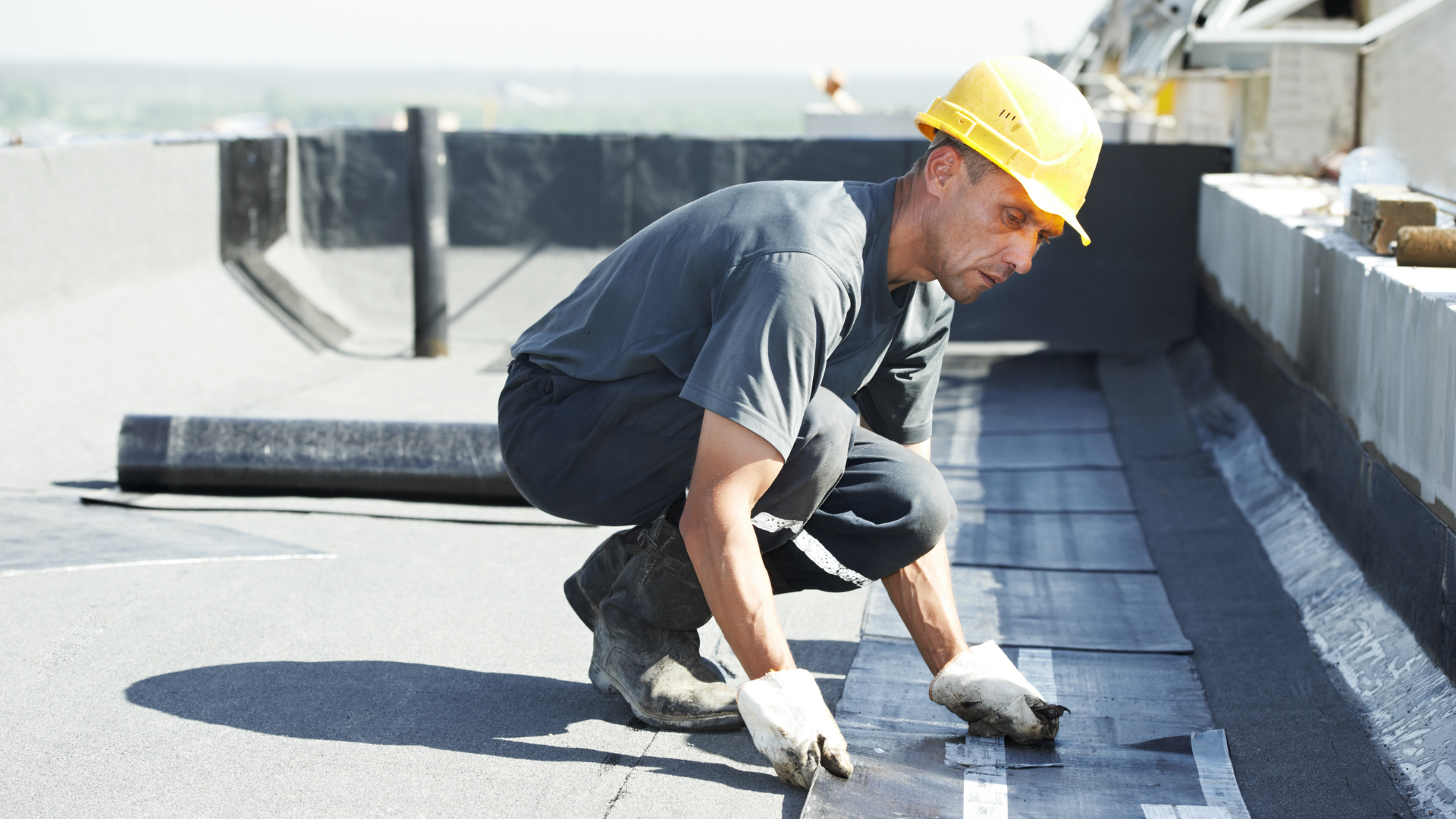 Worker in yellow hard hat applying roofing material on a flat rooftop under bright sunlight