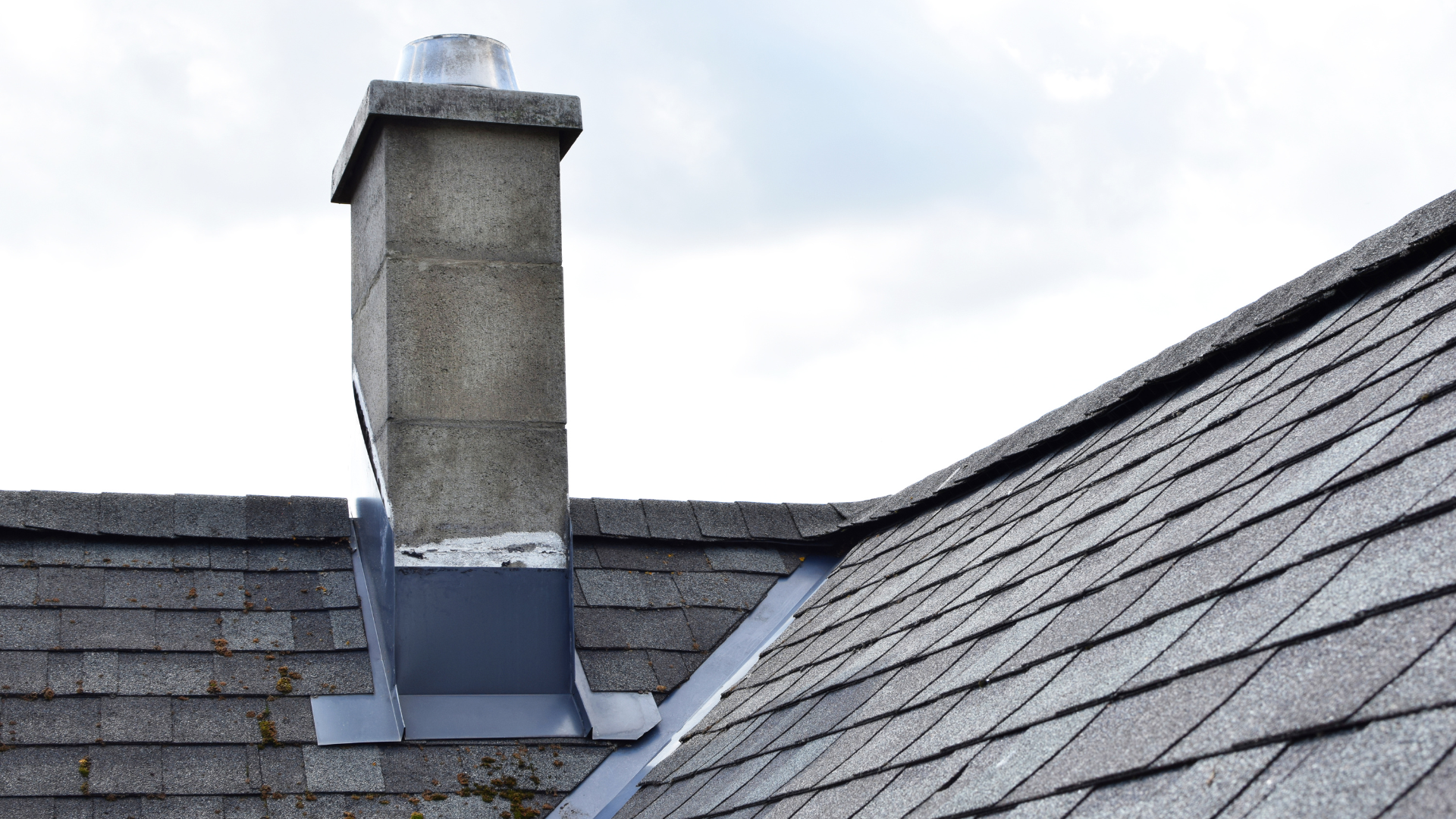 Rooftop chimney with gray stone flue and metal flashing on a shingled roof under a cloudy sky