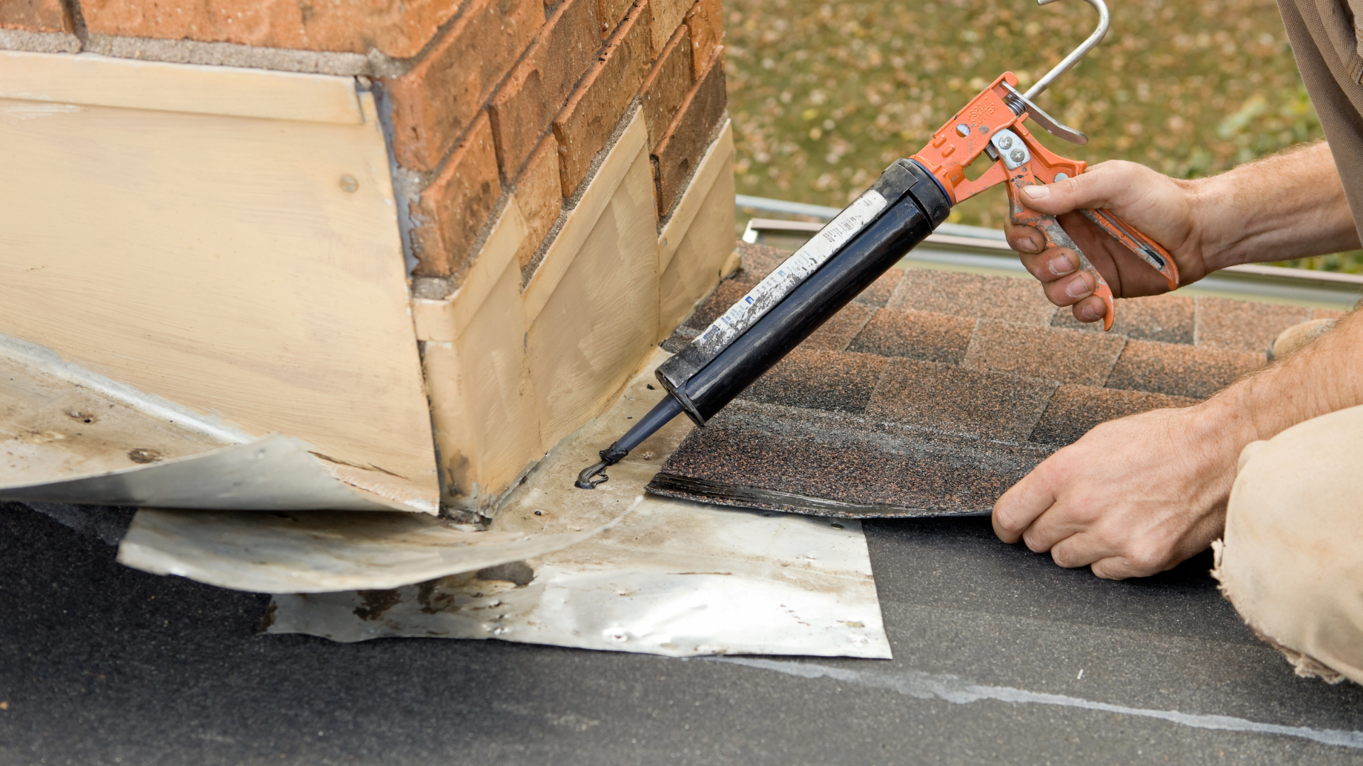 Hand applying roofing sealant with a caulking gun along a flashing on a brick wall base on a flat roof