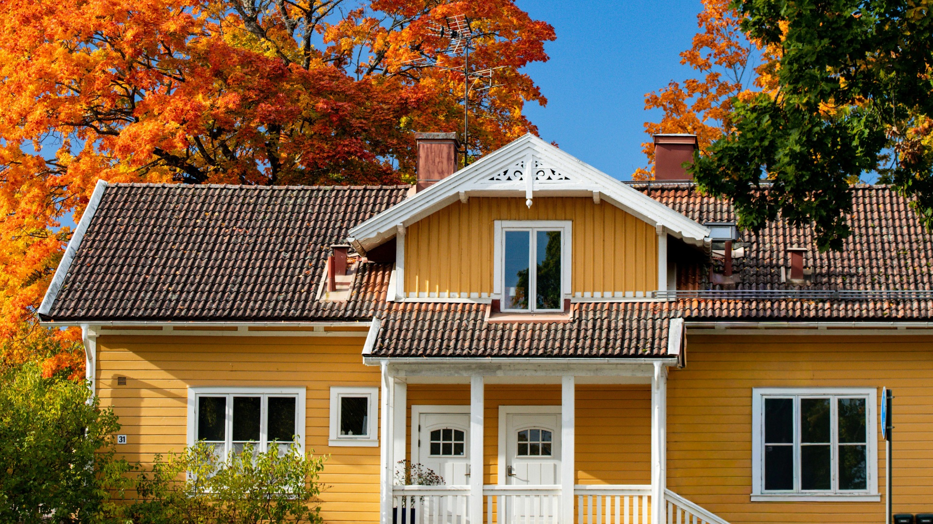 Yellow house with white trim and red-tiled roof, framed by autumn trees with orange leaves.