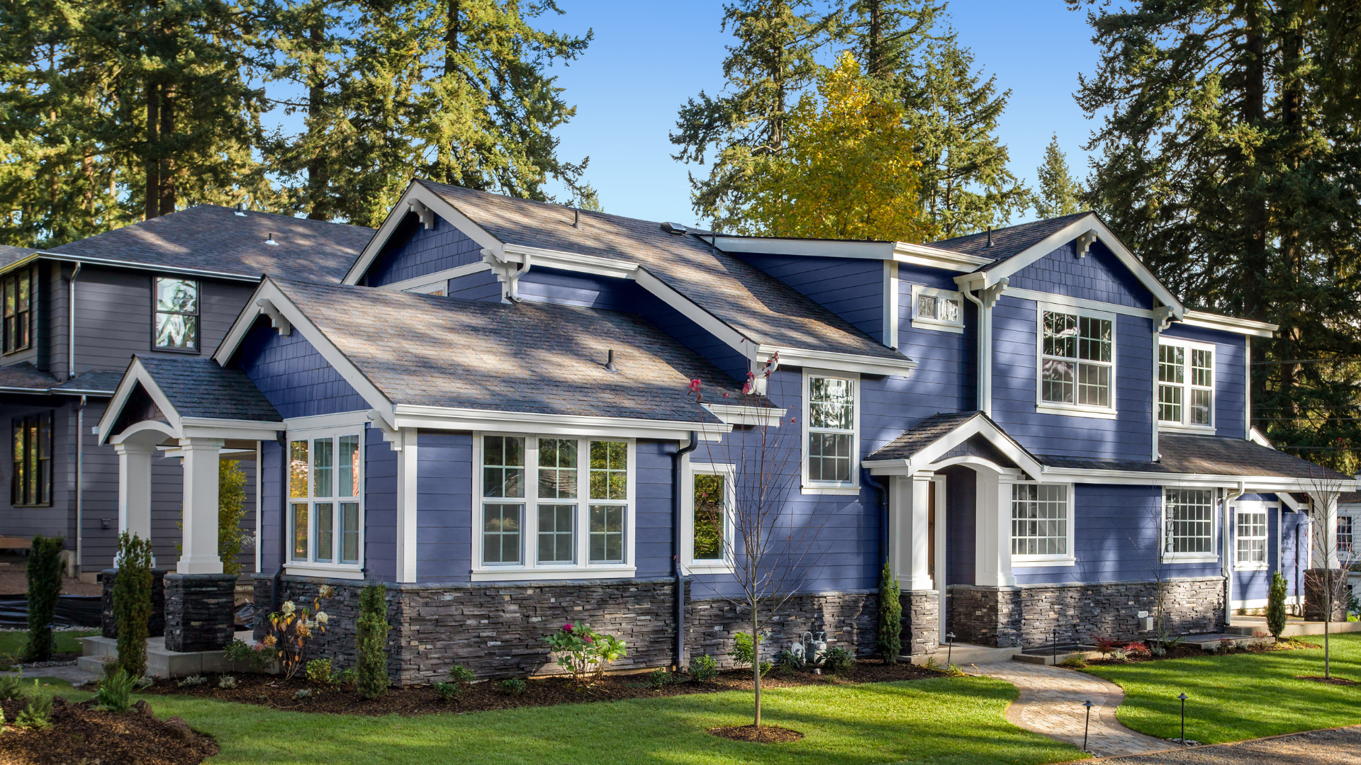 Large blue suburban house with white trim, multiple gables, and tall trees in the yard