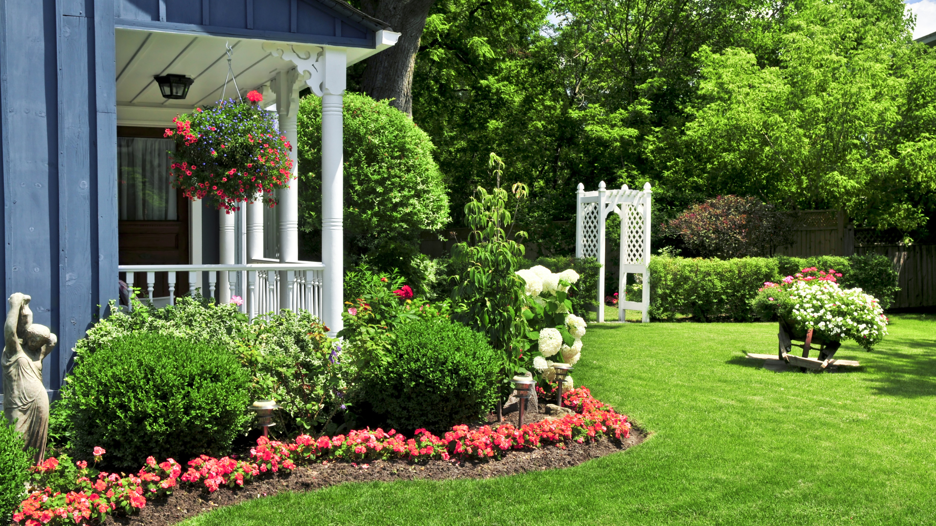 Bright garden with manicured lawn, red flowers, white trellis, and porch of a blue house