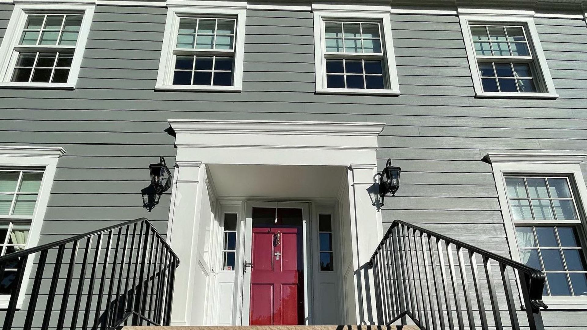 Gray house facade with white trim, black railings, and red front door at the top of steps