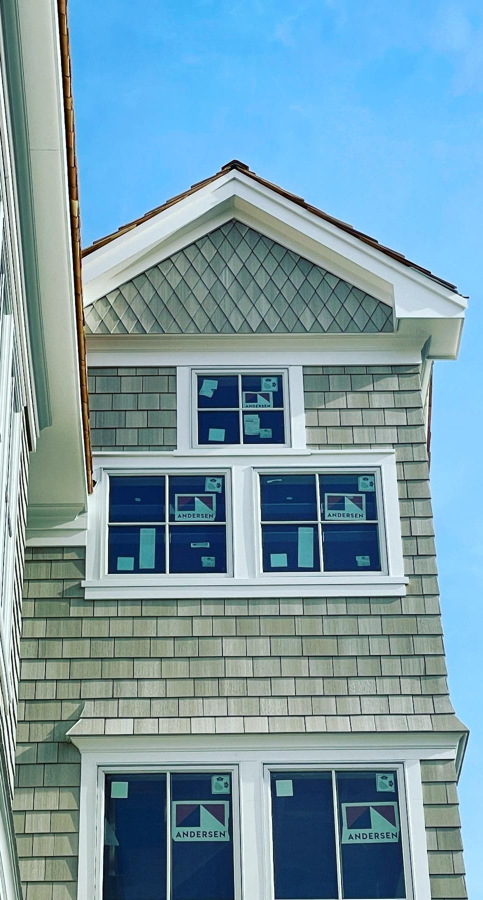 Tall building facade with three sets of windows, shingle siding, and a gabled roof against a blue sky.