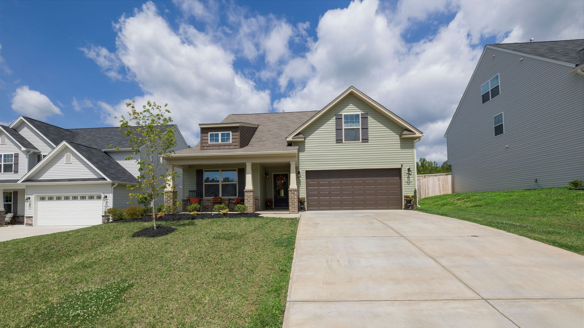 Suburban house with tan siding, brown garage, and a sloped driveway under a partly cloudy sky