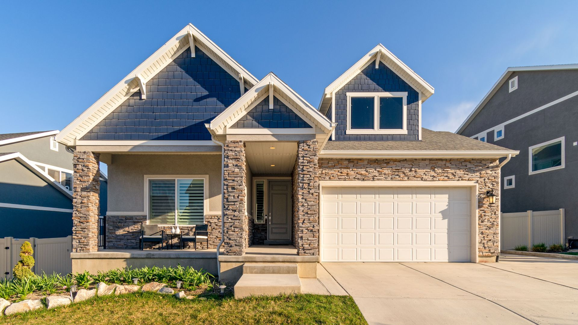 Modern stone-and-blue house with a front porch, dormer windows, and a white garage door under a clear sky