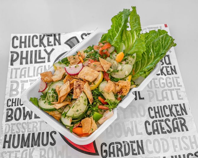Salad in a white container on a food-themed paper, including lettuce, cucumbers, radishes, and pita chips.