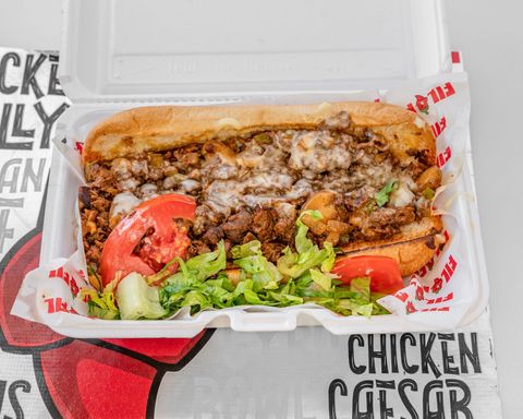 Cheesesteak sandwich with tomatoes and lettuce in a white takeout container.