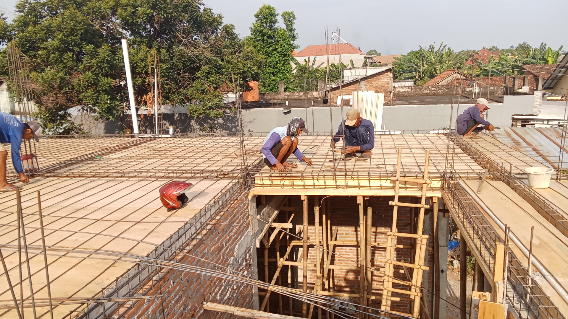 Construction workers on a roof assembling steel reinforcement bars. Wooden forms and surrounding buildings are visible.