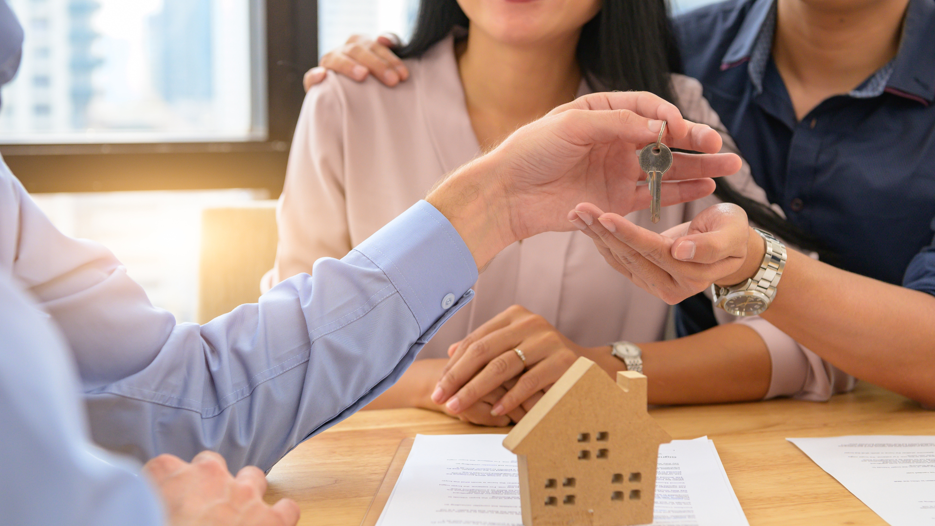 Real estate agent handing keys to a smiling couple, small house model on table.
