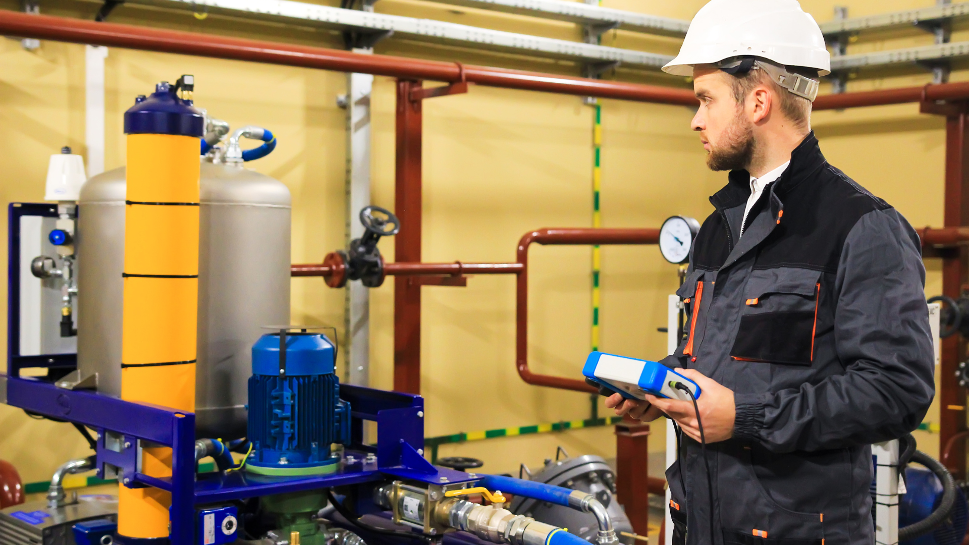 Man in a hard hat inspecting machinery in an industrial setting, holding a device.