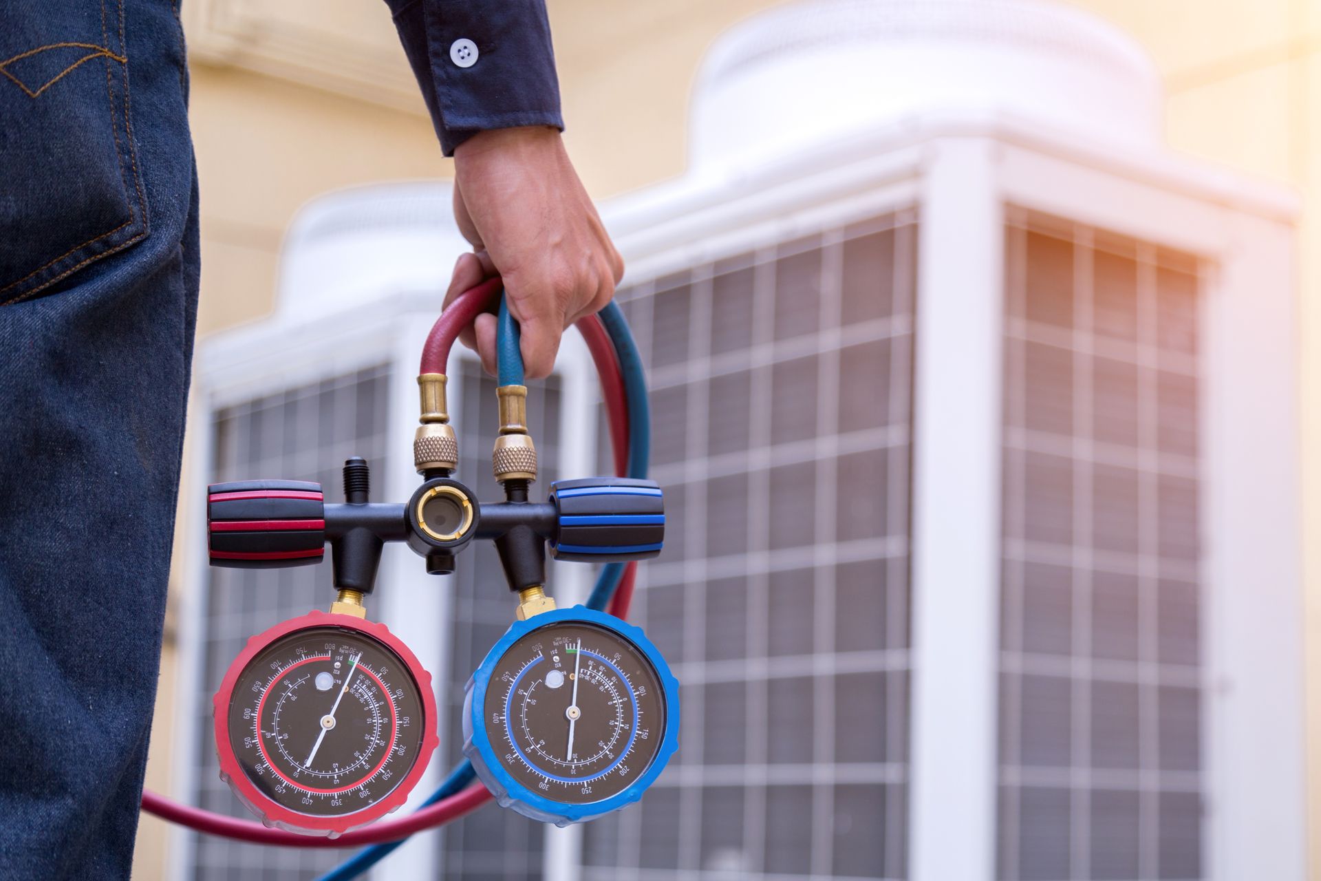 Person holding gauges near an air conditioning unit; blue and red hoses and gauges, outdoors.