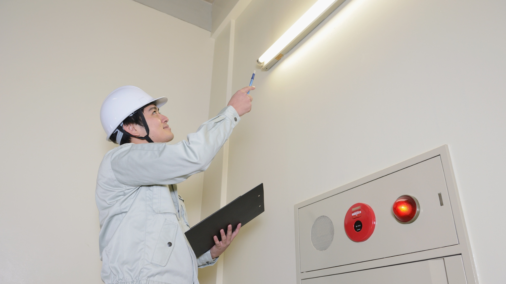 Worker in hard hat inspecting a fluorescent light fixture near a wall control panel.