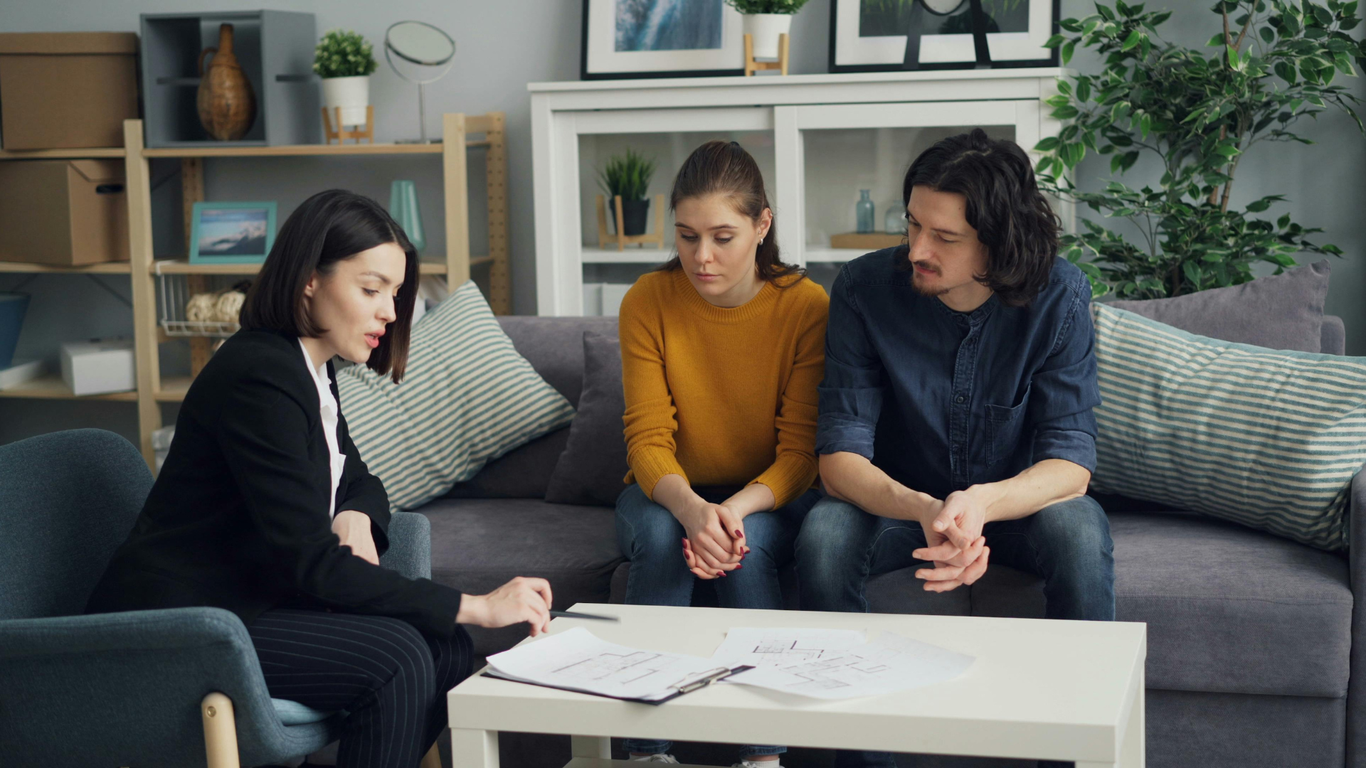A woman in a blazer consults with a couple on a couch, reviewing documents. Indoors.