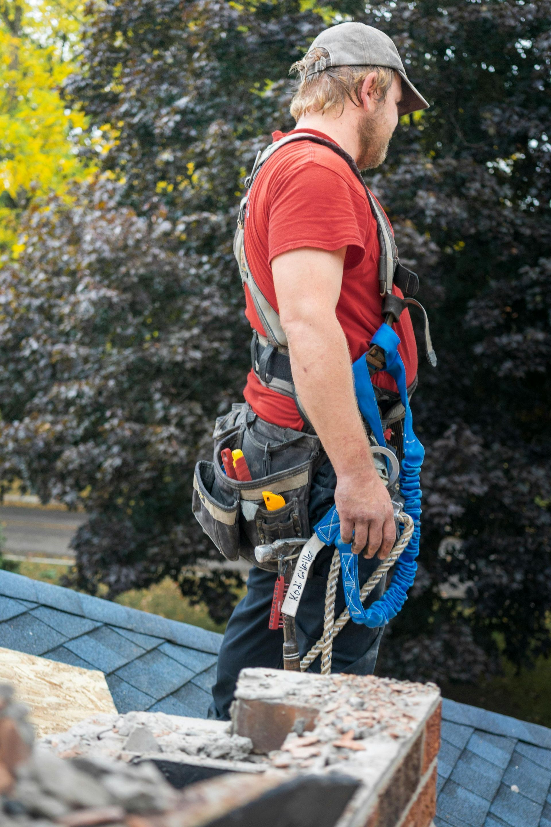 Man on a roof wearing a safety harness, holding a lanyard. Gray cap, red shirt, tool belt, and blue shingles are visible.