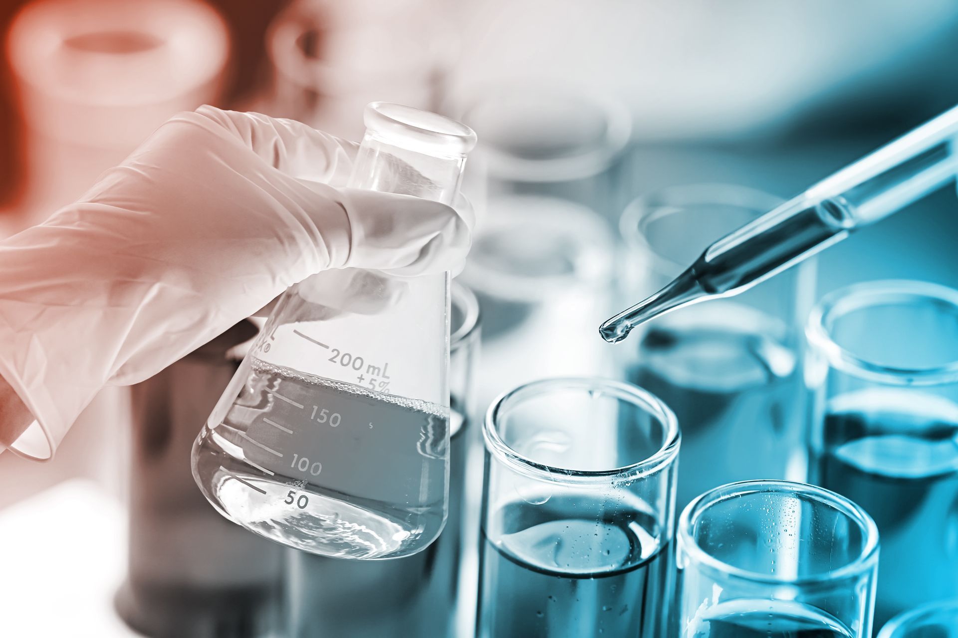 Gloved hand holding a flask, a dropper releasing liquid into a row of test tubes in a lab.