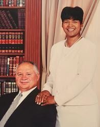 Two people posing indoors, one seated in a suit and one standing in white, smiling by bookshelves and curtains