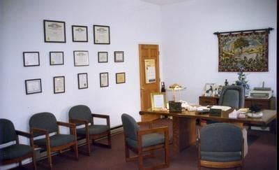 Office waiting room with chairs, framed certificates on the wall, and a desk with a computer and lamp