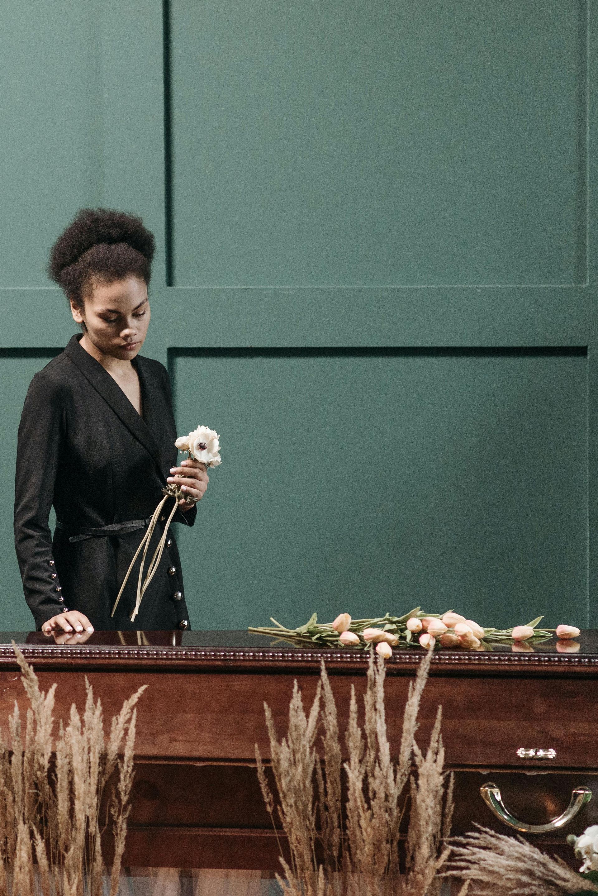 Person in a black dress stands beside a polished wooden casket with flowers in a muted room