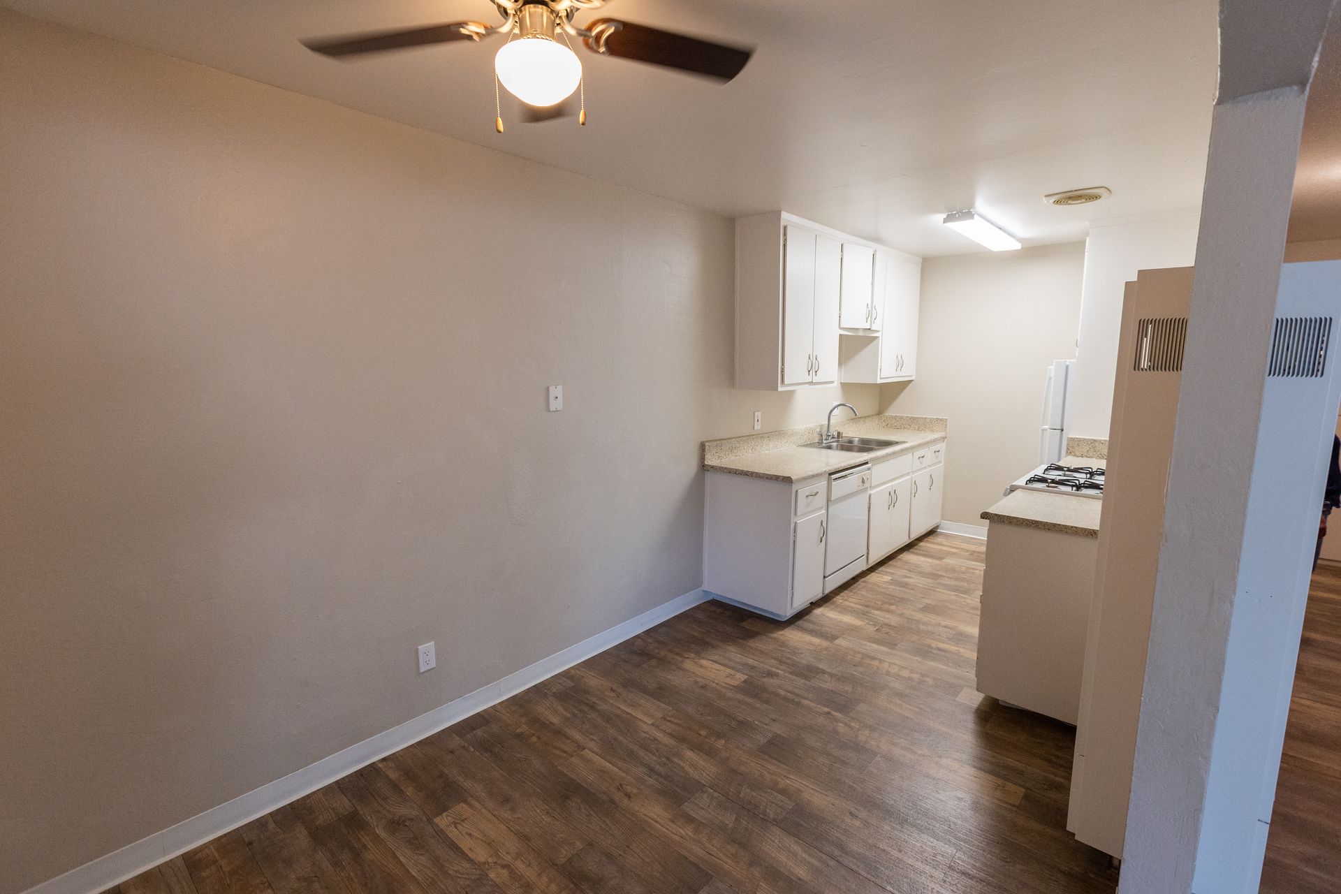 Empty apartment with wood floors, ceiling fan, and small kitchen area.