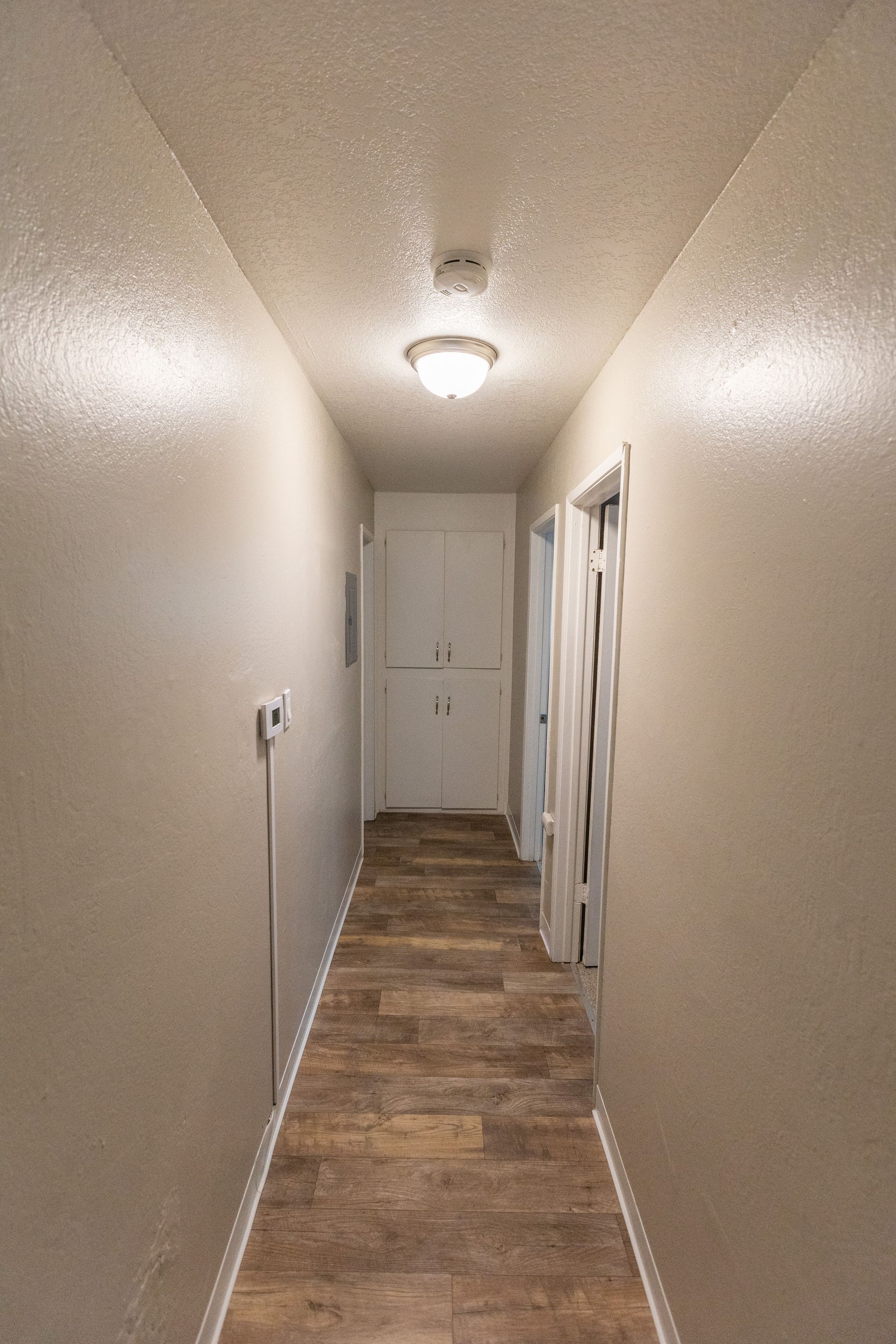 Narrow beige hallway with wood-look flooring, ceiling light, and doors along one side
