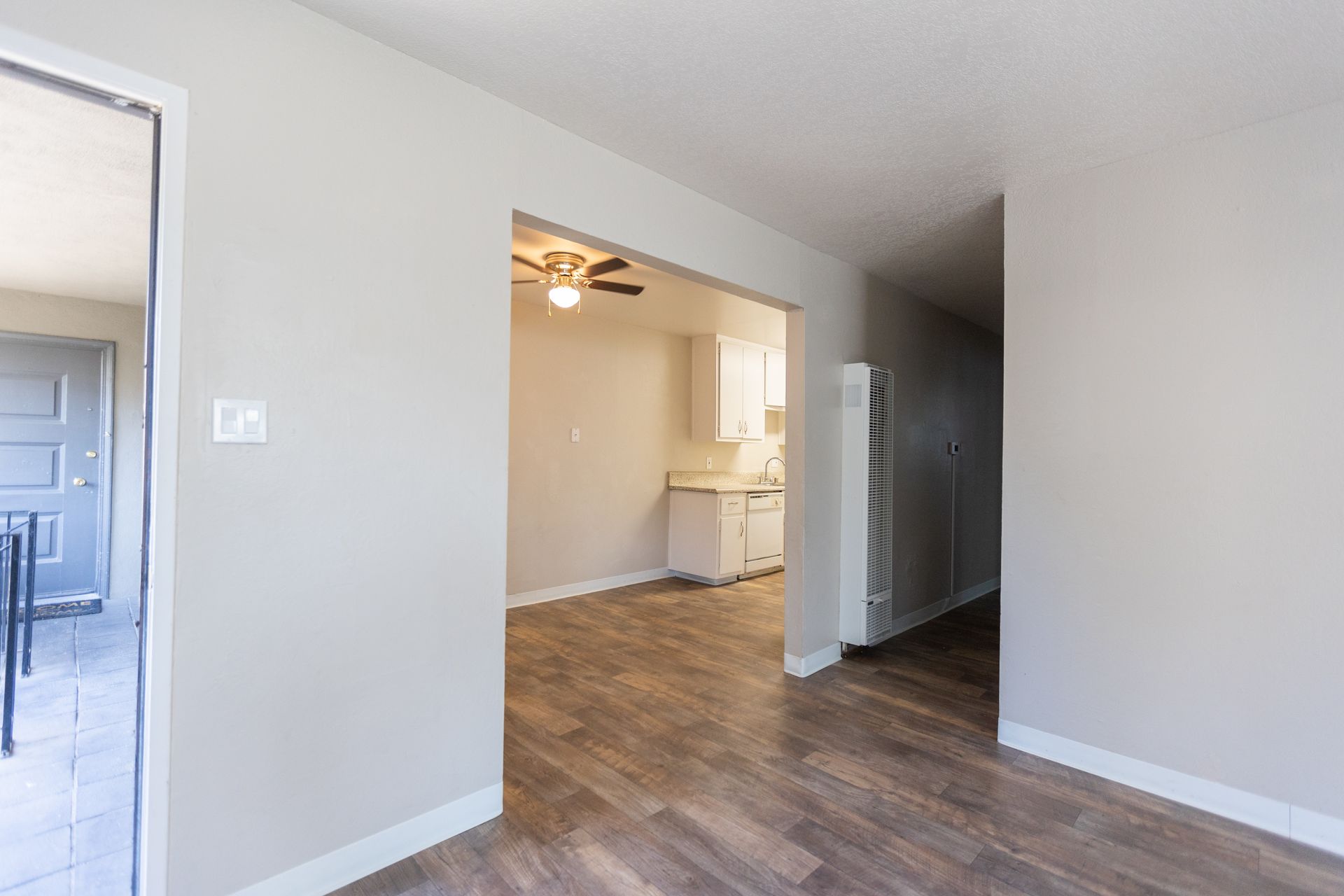 Empty modern room with wood floors, white walls, and a view into a kitchen area