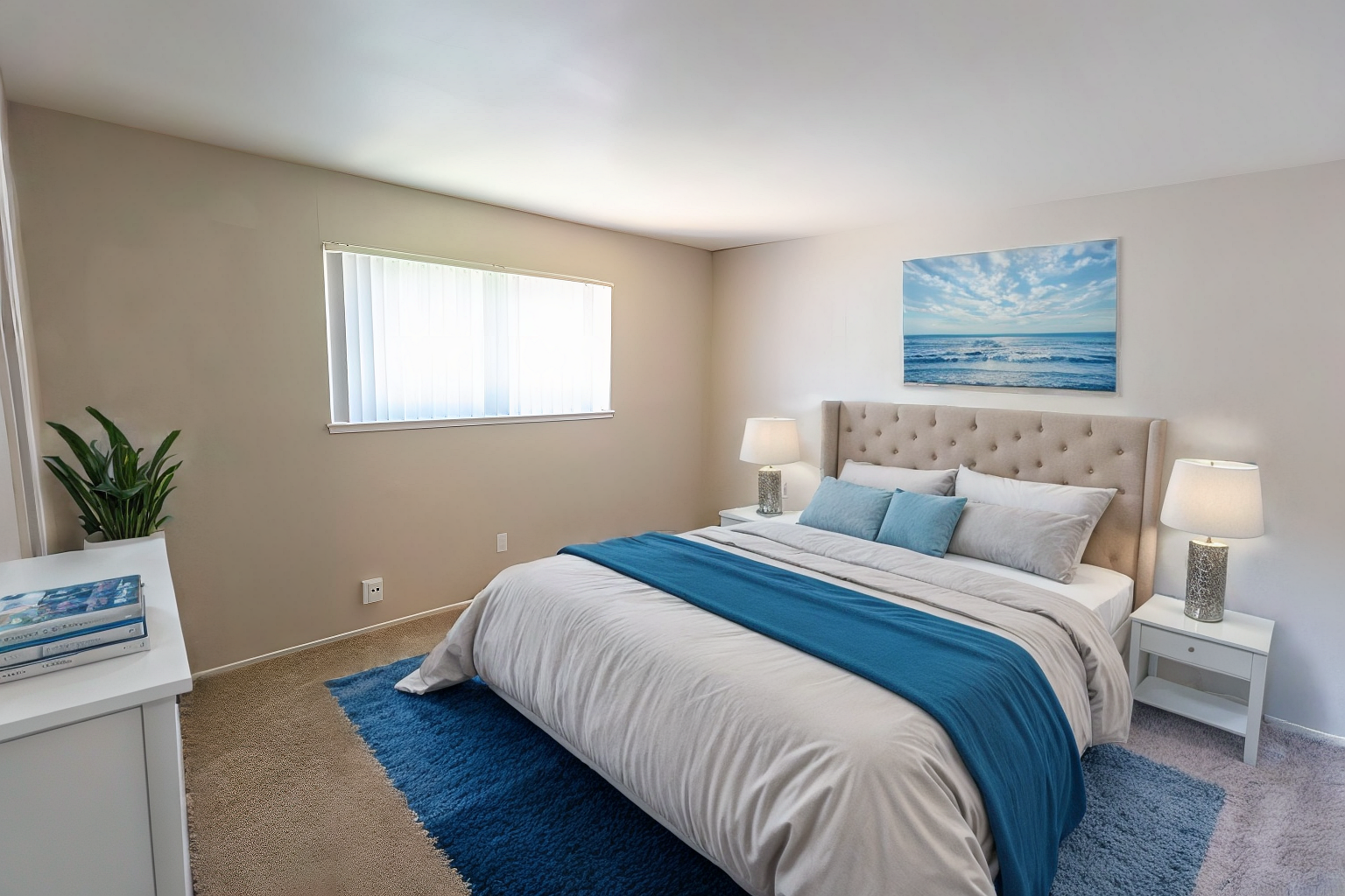 Bright bedroom with a tufted bed, blue accents, window light, and a beach painting above the headboard