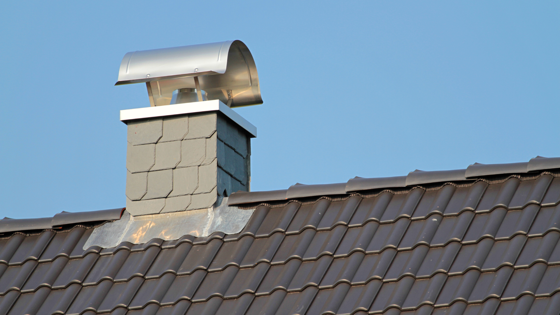Rooftop chimney with silver cap on a dark tiled roof against a clear blue sky
