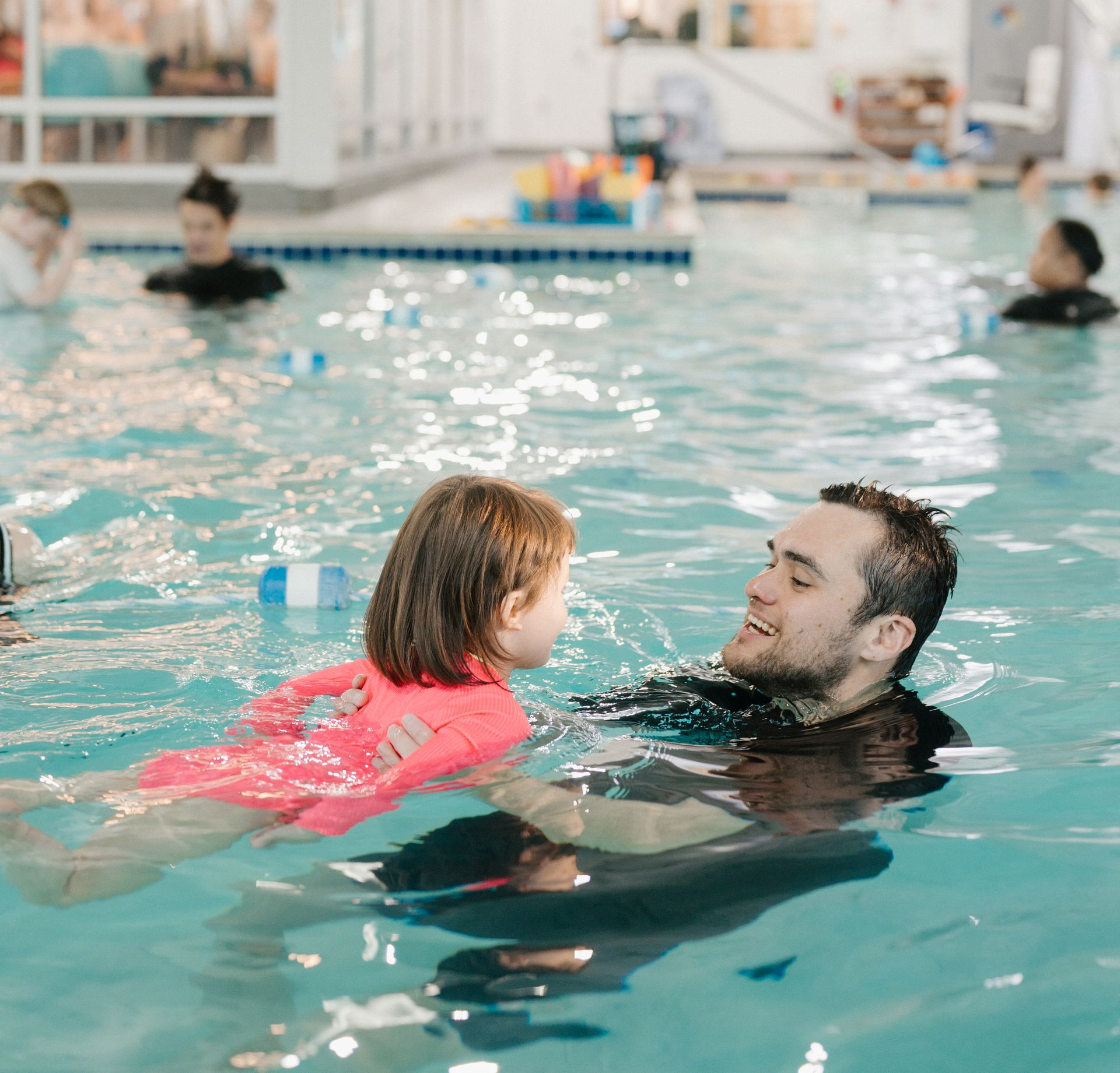 Woman and child in a pool, the child smiling, wearing a red swimsuit, and the woman with an orange hair tie.
