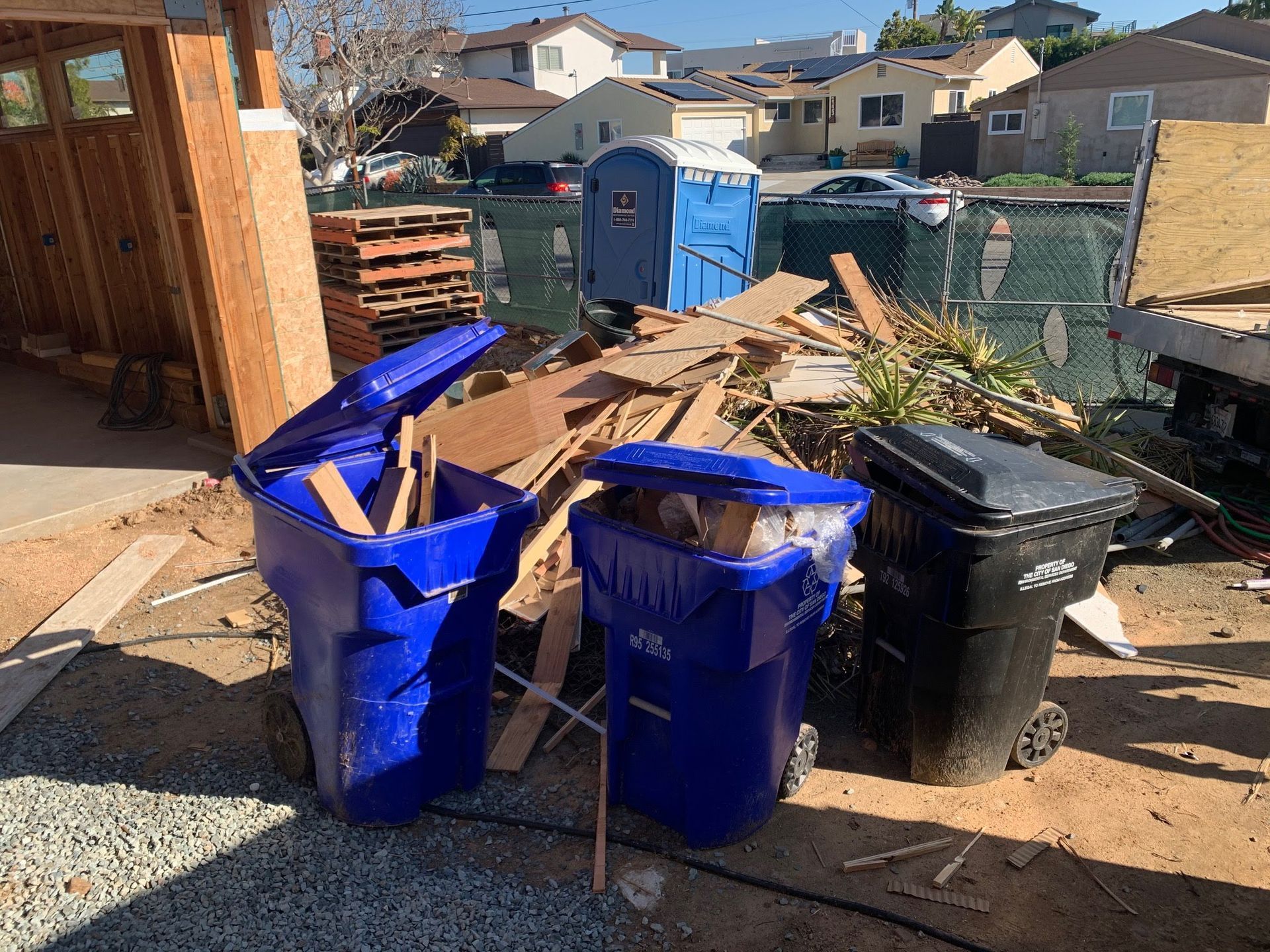 Construction site with two overflowing blue bins and one black bin, debris, and a porta-potty.