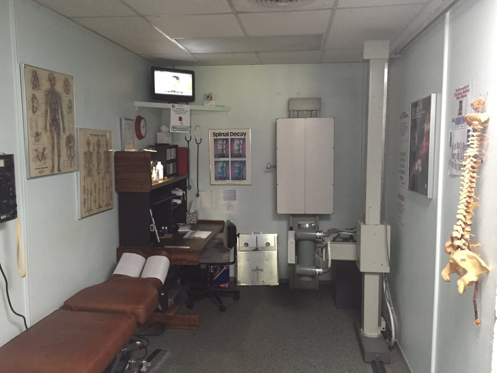 Empty medical exam room with brown exam table, wall diagrams, cabinets, and a sink.