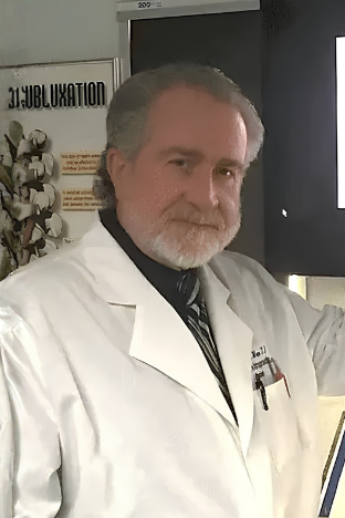 Man in a white lab coat standing in an office doorway, looking at the camera.